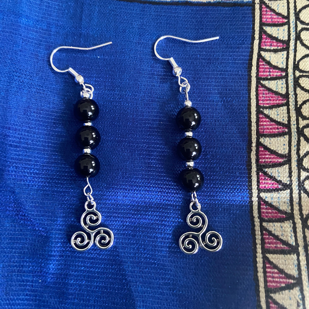 A pair of dangling earrings with Celtic Charms &  3 Onyx or African Turquoise glossy beads, silver bead accents, and silver fishhook ear wires, displayed on a textured royal blue fabric background. 