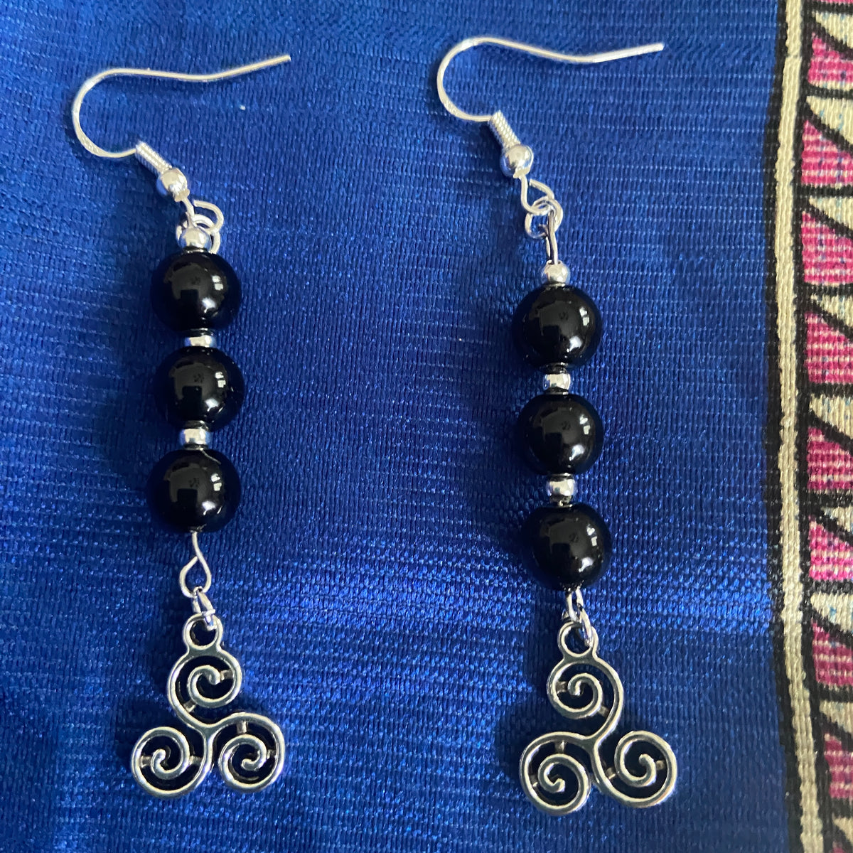 A pair of handmade dangle earrings featuring onyx & African Turquoise  beads with tiny silver beads & Celtic triskelion charms displayed on a blue silk.